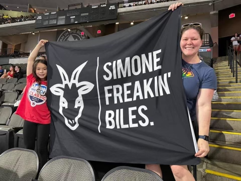 Syd and her mom display a black banner with white lettering and a goat that says Simone Freakin' Biles.